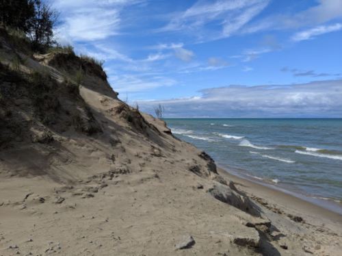 A sandy beach with a steep dune, gentle waves lapping at the shore, and a cloudy blue sky above.