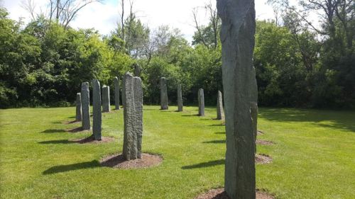A circle of tall stone monoliths surrounded by lush green grass and trees under a bright blue sky.