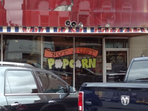 Storefront with a colorful sign reading "Not Just Popcorn," surrounded by parked cars.