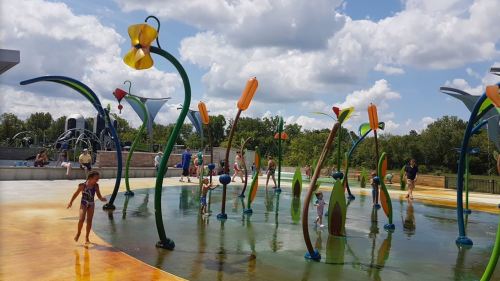 Children play in a colorful splash pad with flower-like water features under a sunny sky.
