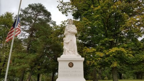 A white statue of a woman holding a child, surrounded by trees, with an American flag nearby.