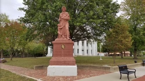 A pink stone memorial statue of a woman holding children, surrounded by trees and grass in a park setting.