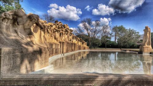A serene outdoor scene featuring a sculptural fountain with figures, surrounded by trees and a clear blue sky.