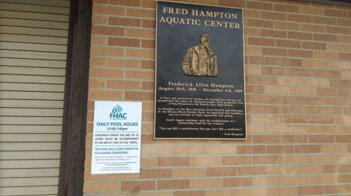 Signage for the Fred Hampton Aquatic Center, featuring a plaque and pool hours notice on a brick wall.