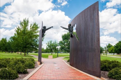 A sculpture of two figures reaching out to each other, framed by large metal panels in a green park setting.