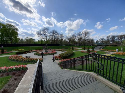 A landscaped garden with colorful flowers, a fountain, and a cloudy blue sky, featuring pathways and greenery.