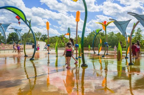 Children play in a colorful splash pad with water features under a bright blue sky and fluffy clouds.