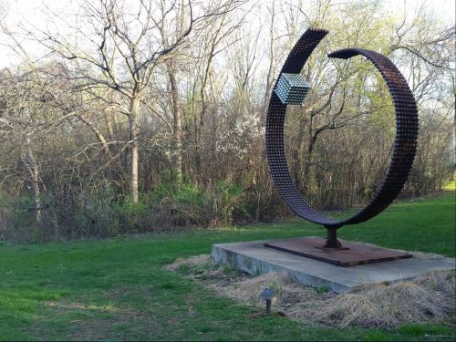 A large, abstract metal sculpture stands on a concrete base in a grassy area, surrounded by trees and greenery.