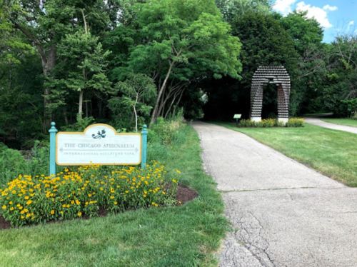 A pathway leads through greenery, with a sign for the Chicago Athenaeum and a sculpture archway in the distance.