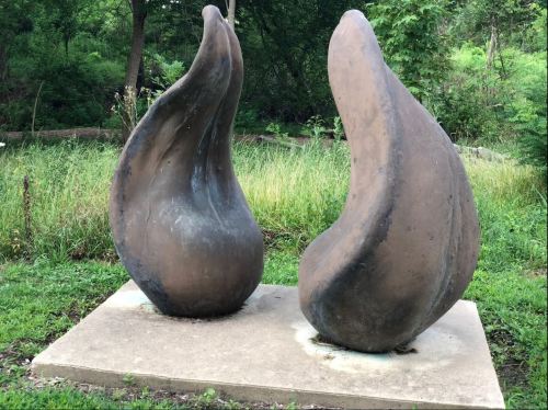 Two abstract, curved sculptures on a concrete base, surrounded by lush greenery in a natural setting.
