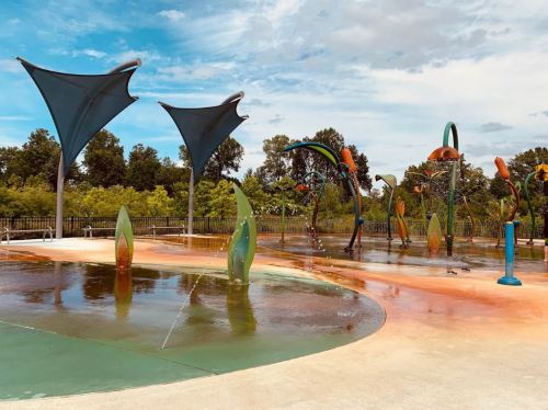 Colorful splash pad with water features and large shade structures, surrounded by greenery under a blue sky.