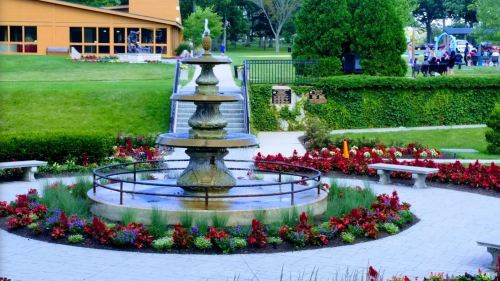 A decorative fountain surrounded by colorful flowers in a park setting, with green grass and trees in the background.