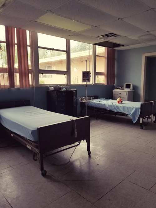 Two empty hospital beds in a room with large windows, a medical monitor, and minimal furnishings.