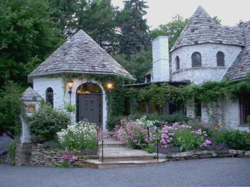 Charming stone cottage with a turret, surrounded by colorful flowers and lush greenery. Warm evening light glows from the entrance.