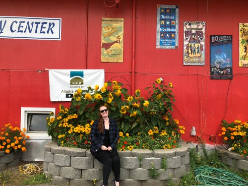 A woman sits on a stone planter surrounded by colorful flowers in front of a red wall with posters and a banner.