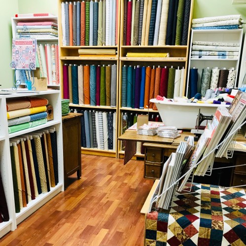 A cozy fabric store with colorful rolls of fabric on shelves and a quilting table in the foreground.