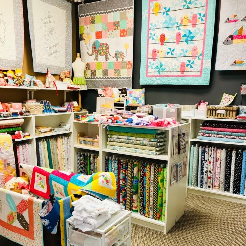 A cozy fabric shop filled with colorful quilts, fabrics, and sewing supplies displayed on shelves and tables.