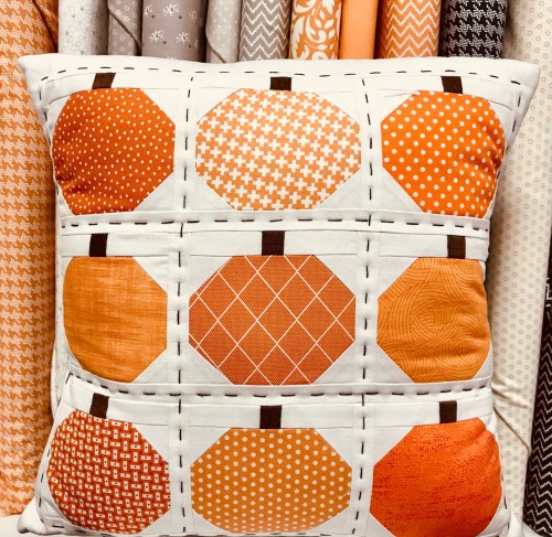 A decorative pillow featuring a patchwork design of orange pumpkins on a white background, surrounded by fabric swatches.