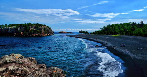 A scenic view of a black sand beach with gentle waves, surrounded by lush greenery and rocky formations under a blue sky.