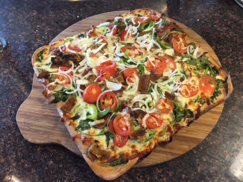 A square pizza topped with cheese, tomatoes, onions, and greens on a wooden cutting board.