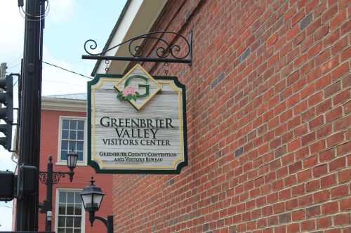 Sign for the Greenbrier Valley Visitors Center hanging from a post, with a brick building in the background.