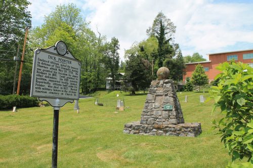 A stone monument with a globe on top, next to a historical marker, set in a grassy area with trees and a building in the background.