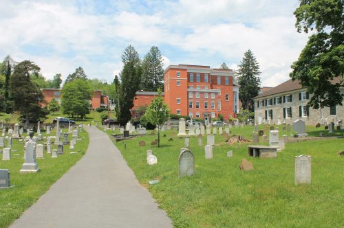 A peaceful cemetery with gravestones, a paved path, and buildings in the background under a partly cloudy sky.