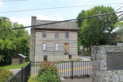 Historic log cabin with a stone foundation, surrounded by greenery and a road, featuring a wooden door and nearby signage.