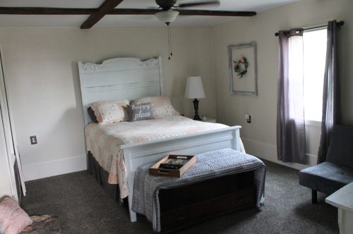 Cozy bedroom with a white bed, decorative pillows, a lamp, and a gray chair, featuring soft lighting and neutral decor.