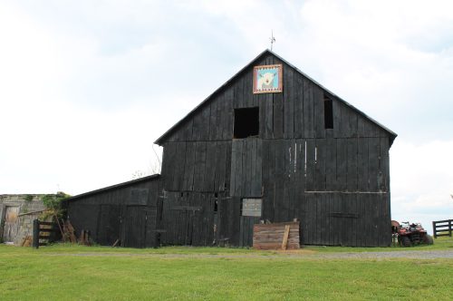 A weathered black barn with a decorative sign, surrounded by green grass and a cloudy sky.
