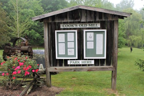 A wooden sign for Cook's Old Mill with informational panels, surrounded by greenery and flowers.