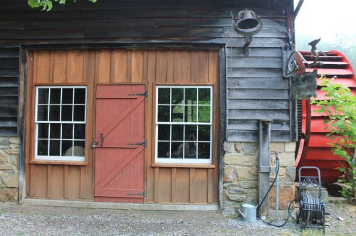A rustic wooden building with a red water wheel, featuring two windows and a brown door.