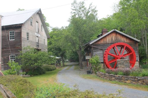 A gravel path leads between two rustic buildings, featuring a large red water wheel beside a stone wall.