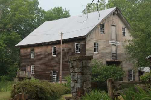 A rustic wooden mill building surrounded by greenery, featuring a metal roof and large windows.