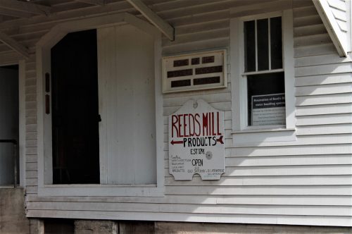 Sign for Reeds Mill Products on a white wooden building, indicating it's open and providing historical information.