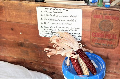 A sign listing product information next to a bundle of dried corn and a blue bucket in a rustic setting.