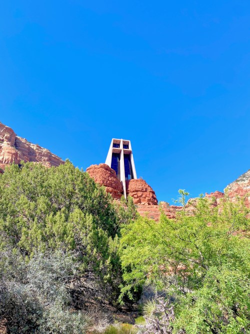 A tall structure stands atop red rock formations under a clear blue sky, surrounded by green vegetation.
