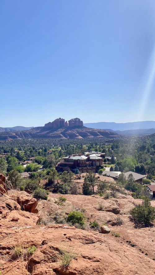 A scenic view of red rock formations and green trees under a clear blue sky, with buildings nestled in the foreground.