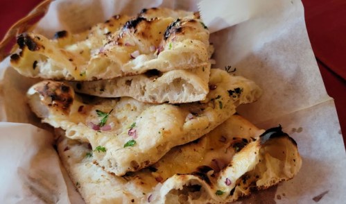 A stack of freshly baked naan bread, lightly charred and garnished with herbs and onions, served in a basket.