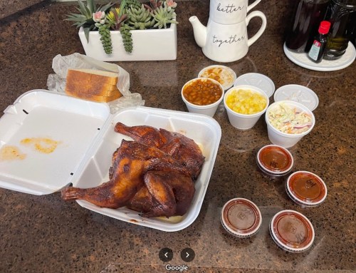 A takeout meal featuring smoked chicken, sides of baked beans, coleslaw, corn, and sauces, with a slice of bread.
