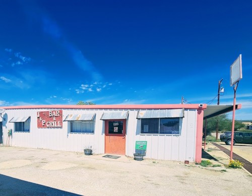A small, rustic bar and grill building with a red sign, surrounded by a clear blue sky and gravel parking area.