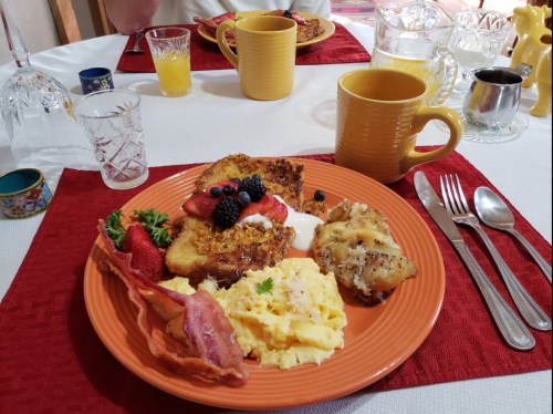 A colorful breakfast plate with French toast, berries, scrambled eggs, bacon, and a drink on a table setting.