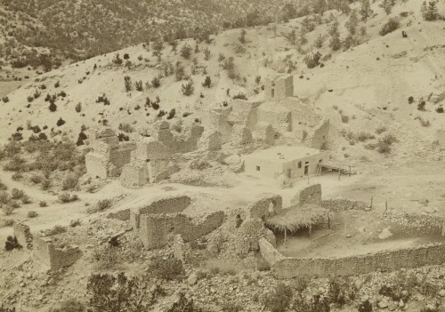 Black and white image of ancient stone ruins in a desert landscape, surrounded by sparse vegetation and hills.