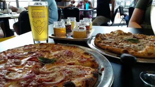 A table with two pizzas, a flight of beer samples, and a glass of beer in a lively restaurant setting.