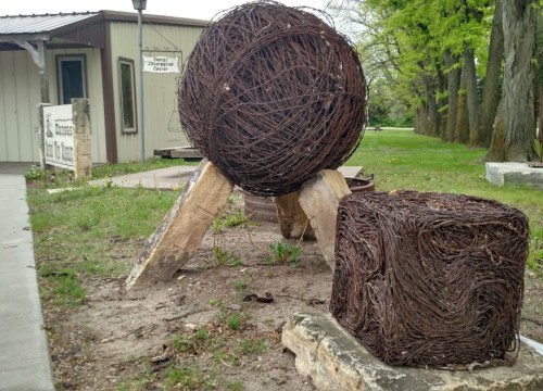 A large spherical sculpture and a smaller cube, both made of intertwined twigs, set in a grassy area near a building.