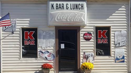 Exterior of K Bar & Lunch featuring a Coca-Cola sign, window decals, and flower pots at the entrance.