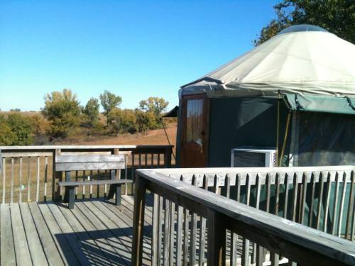 A wooden deck with a bench overlooks a grassy field and a yurt under a clear blue sky.
