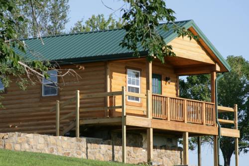 A wooden cabin with a green metal roof, featuring a porch and stone foundation, surrounded by trees and blue sky.