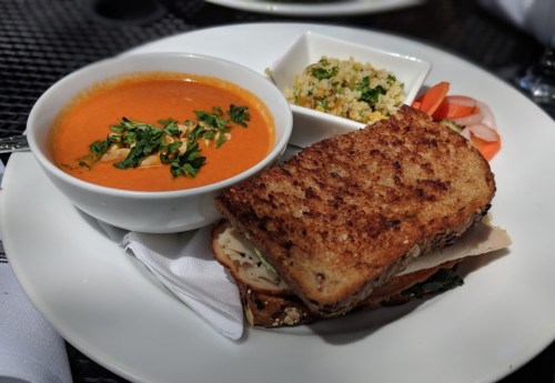 A plate with a grilled sandwich, tomato soup, a side of couscous, and sliced vegetables garnished with herbs.