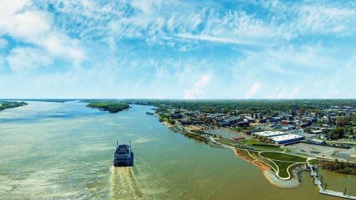Aerial view of a river with a ferry, lush green banks, and a city skyline under a bright blue sky.